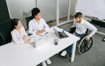 Three people sat around a desk, in an office. One person is in a wheelchair.