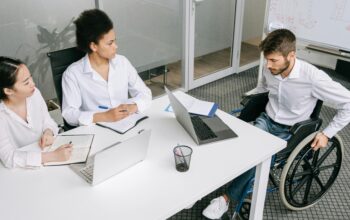 Three people sat around a desk, in an office. One person is in a wheelchair.