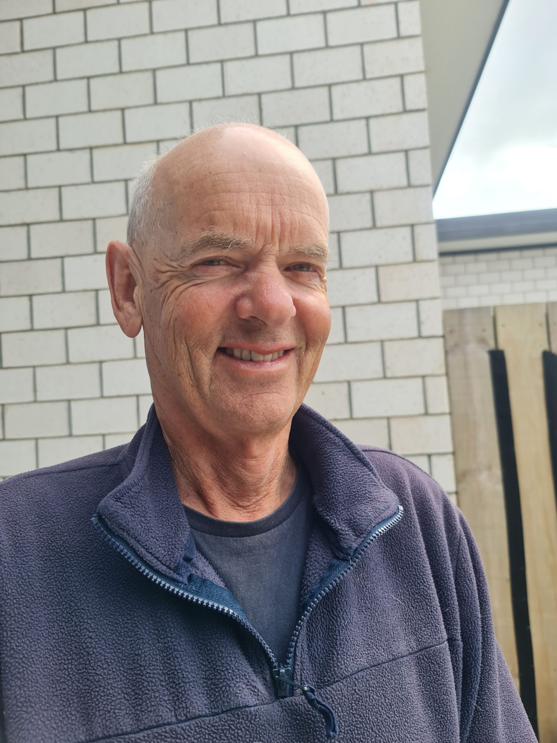 A person smiling outside, with a brick wall background.