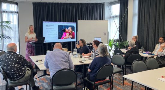 People are sat round a table while a woman at the front of the room delivers a workshop and presentation on Disability Support.